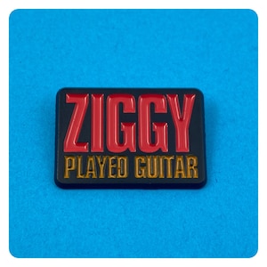 May include: A black enamel pin with the text "ZIGGY PLAYED GUITAR" in red and gold letters.