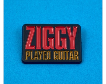 May include: A black enamel pin with the text "ZIGGY PLAYED GUITAR" in red and gold letters.