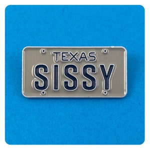 Könnte beinhalten: Rechteckiger Ansteck-Pin, der einem Nummernschild aus Texas ähnelt. Der Pin ist hellgrau mit dem Wort "TEXAS" oben und dem Wort "SISSY" in fetten, dunkelblauen Buchstaben. Er hat einen silbernen Rand und vier kleine silberne Nieten.