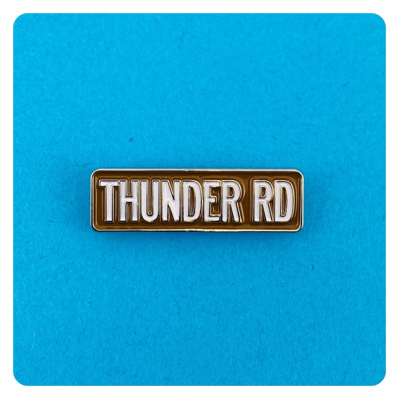 May include: A brown enamel pin with a silver border. The pin reads "THUNDER RD" in white letters.