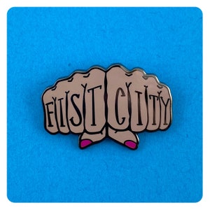 May include: A metal pin with two fists forming the word "FIST CITY". The fists are a light brown color with pink fingernails. The background is a solid blue.