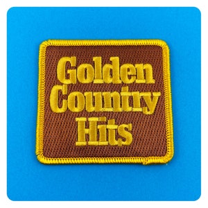 May include: A brown fabric patch with yellow stitching. The patch has the text "Golden Country Hits" embroidered in yellow thread.