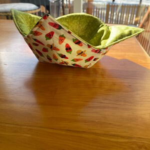 May include: A green and white fabric bowl with a pattern of red peppers and butterflies. The bowl is folded and sitting on a wooden table.