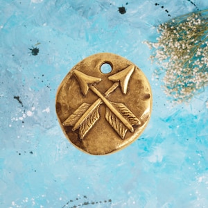 May include: A gold-toned metal charm with a design of two crossed arrows. The charm has a slightly textured surface and a hole at the top for attaching to a necklace or bracelet.