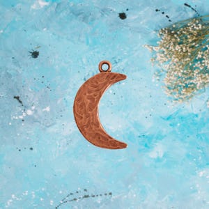 May include: A copper crescent moon charm with a textured surface. The charm has a loop at the top for attaching to a necklace or bracelet.