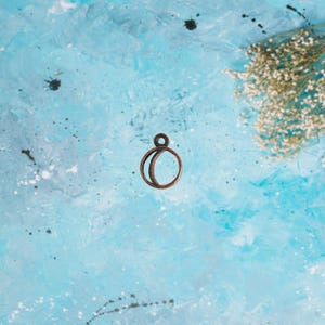 May include: A bronze-colored charm with a circular design, featuring a smaller circle attached to a larger, partially overlapping circle. The charm is set against a textured, light blue background with white accents and a small cluster of dried flowers.