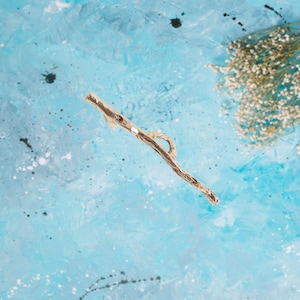 May include: A gold-colored, twig-shaped decorative object with a loop and a small, round accent. The object is set against a light blue, textured background with white and black paint splatters and a cluster of dried flowers.