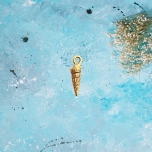 May include: A gold-colored seashell charm with a loop at the top. The charm is on a blue background.