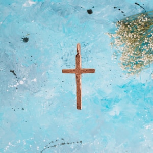 May include: A hammered copper cross pendant with a rustic finish. The pendant is hanging against a light blue background.
