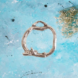 May include: A silver-colored, open-circle bracelet with a textured, organic design. The bracelet has a branch-like appearance, with a loop at the top and a textured base. The background is a blue and white abstract design.
