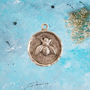 May include: Silver charm with a bee design. The bee is in a raised, textured design. The charm is round and has a loop at the top for attaching to a necklace or bracelet.