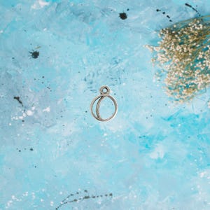 May include: A silver-toned charm with a crescent moon design, featuring a small loop at the top for attachment. The charm is set against a textured blue background with white accents and dried flowers.