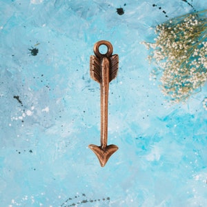 May include: A copper arrow charm with a loop at the top for attaching to jewelry or other crafts. The arrow is pointing to the right.