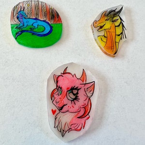 May include: Three 2D plastic figurines. A blue dragon with a green background, a yellow dragon with an orange background, and a pink furry creature with a red heart.