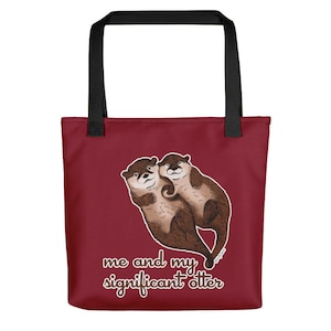 Puede incluir: Una bolsa de tela de color granate con asas negras presenta una ilustración de dibujos animados de dos nutrias abrazándose. El texto "me and my significant otter" está impreso en marrón debajo de las nutrias.