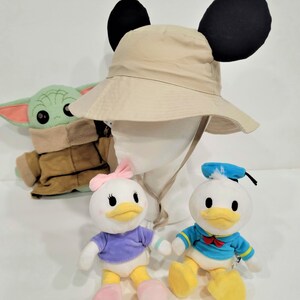 May include: A beige bucket hat with black Mickey Mouse ears, a green and brown plush toy, a white plush duck with a purple shirt and pink bow, and a white plush duck with a blue shirt and blue sailor hat.
