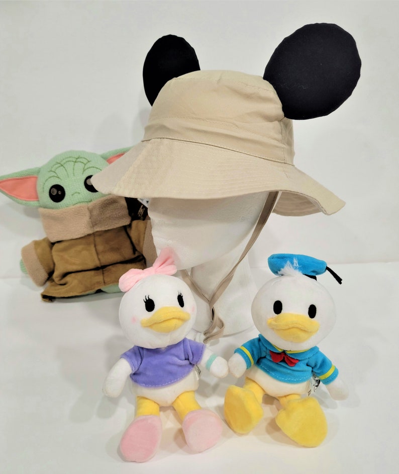 May include: A beige bucket hat with black Mickey Mouse ears, a green and brown plush toy, a white plush duck with a purple shirt and pink bow, and a white plush duck with a blue shirt and blue sailor hat.