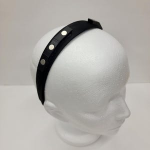 May include: A black headband with silver metal accents. The headband is on a white mannequin head.