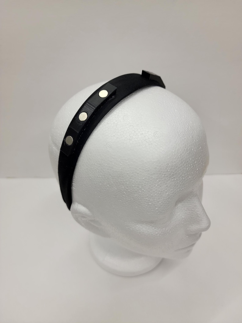 May include: A black headband with silver metal accents. The headband is on a white mannequin head.
