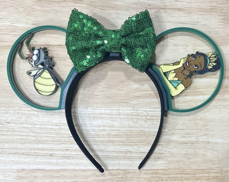 May include: Green and black mouse ears with a green bow, a cartoon frog and a cartoon princess in a green dress.