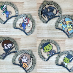 Op de afbeelding: Houten uitsparingen van personages met Luigi, Mario, Toothless, een draak, Harry Potter, Hermelien, een stripfiguur en het monster van Frankenstein. Elk personage is geplaatst in een decoratief, cirkelvormig frame met een goudkleurige bovenkant.