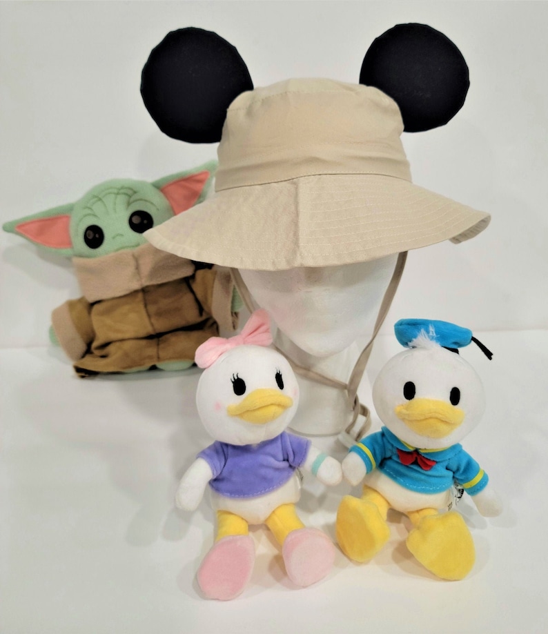 May include: A beige Mickey Mouse hat with black ears, a green and brown plush Baby Yoda, and two plush Disney ducks, one with a blue sailor hat and the other with a pink bow.