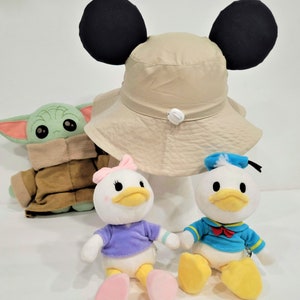May include: A beige bucket hat with black Mickey Mouse ears, a green plush Baby Yoda, a white plush Daisy Duck with a purple shirt and pink bow, and a white plush Donald Duck with a blue sailor shirt.
