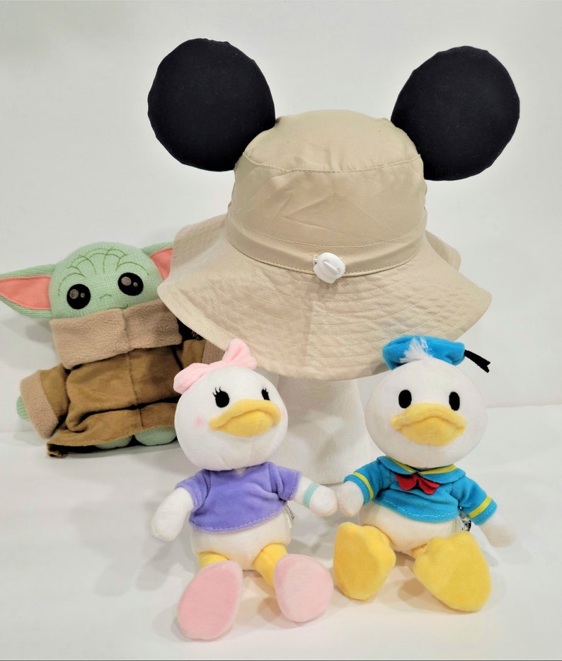 May include: A beige bucket hat with black Mickey Mouse ears, a green plush Baby Yoda, a white plush Daisy Duck with a purple shirt and pink bow, and a white plush Donald Duck with a blue sailor shirt.