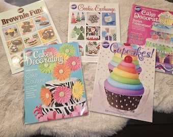 May include: Five Wilton instructional books featuring colorful photography of baked goods. Titles include Brownie Fun!, Cookie Exchange, Cake Decorating Sweet Celebrations, Cake Decorating Cake Couture!, and Cupcakes! with 320 designs.