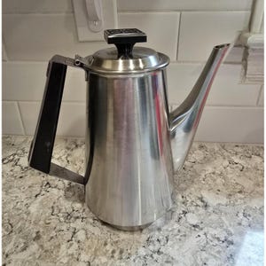 May include: A stainless steel coffee pot with a tall, tapered body, a long angled spout, and a black handle. The lid features a black square knob with a decorative pattern on top. The metal surface is polished and reflective with a smooth finish.