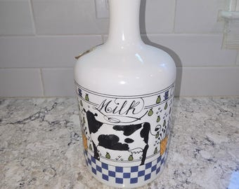 May include: A white milk bottle with a narrow neck and a flared rim. The body features a black and white cow illustration, the word Milk in cursive script, green pear accents, and a blue and white checkered pattern along the bottom edge.