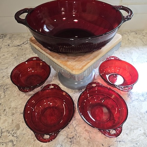 May include: A set of ruby red glass serving dishes. The set includes a large bowl with handles and four smaller oval dishes. The dishes have a ribbed design and are arranged on a countertop.
