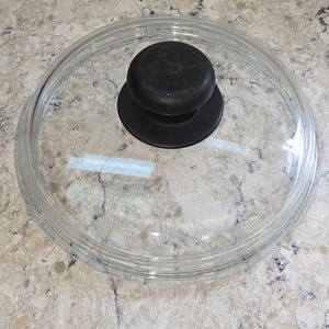 May include: A circular, transparent glass pot lid featuring a smooth, rounded black knob handle centered on top. The lid has a raised outer rim designed to fit securely onto a cooking vessel, providing a clear view of the contents inside.