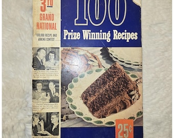 May include: A vintage Pillsbury 3rd Grand National 100 Prize Winning Recipes cookbook with a blue cover. The front features a slice of chocolate cake on a leaf-patterned plate, black and white photos of contest winners, and a 25 cent price tag.