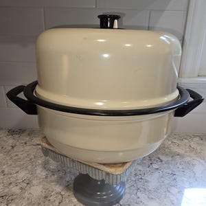 May include: Cream-colored vintage oven with a black rim and handles. The oven has a matching lid with a black handle. It sits on a decorative, gray and silver cake stand.