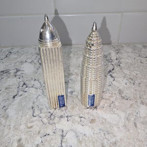 May include: Two silver-plated salt and pepper shakers. One is a tall, rectangular shape with vertical ridges. The other is a skyscraper design. Both have a pointed top and a blue label.
