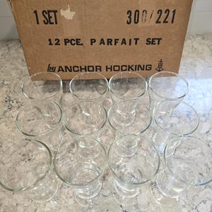 May include: A set of twelve clear glass parfait glasses arranged in front of a cardboard box. The box is labeled "1 SET" and "12 PCE. PARFAIT SET" with the Anchor Hocking logo.