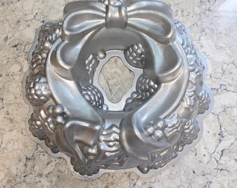 May include: A silver-colored metal bundt cake pan featuring a molded design of a decorative bow at the top, surrounded by clusters of berries and leaves. The pan has a circular center hole and a scalloped edge, with a smooth, metallic finish.