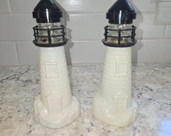 May include: A pair of lighthouse-shaped salt and pepper shakers featuring a white, brick-textured body with a molded door and window. Each shaker is topped with a black lantern room, a black railing, and a clear glass section for the seasoning.