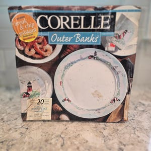 May include: A Corelle Outer Banks 20-piece dinnerware set box featuring a nautical theme with lighthouse and sailboat illustrations. The packaging displays a bowl of shrimp, a plate with a lighthouse design, and a break and chip resistant label.