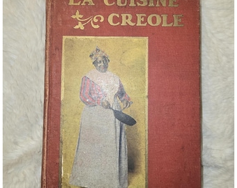 May include: A red hardcover book titled LA CUISINE CREOLE in gold lettering. The cover features a rectangular illustration of a person wearing a head covering, a striped long-sleeve shirt, and a long skirt, holding a black frying pan in one hand.