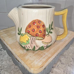 May include: Cream-colored ceramic pitcher with a whimsical mushroom design. The pitcher features a brown mushroom with red spots, green leaves, and a yellow handle. The pitcher is sitting on a wooden tray with a gray scalloped edge.