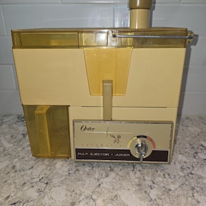 May include: Vintage Oster automatic pulp ejector juicer in cream and tan. The juicer has a control panel with a dial and the Oster logo. The juicer is made of plastic and has a rectangular shape.
