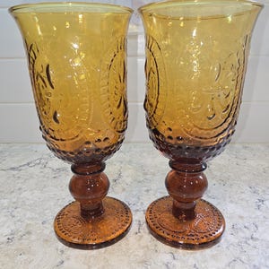 May include: Two amber-colored vintage goblets. The glasses have a decorative pattern on the bowl and base. The stem is a dark brown color with a round ball in the middle. The base is round with a decorative pattern.