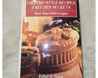 May include: A cookbook titled Mennonite Country-Style Recipes & Kitchen Secrets by Esther H. Shank. The cover features a large, golden-brown baked pie with a fluted crust, set on a stone surface, with text stating More than 1,000 recipes.