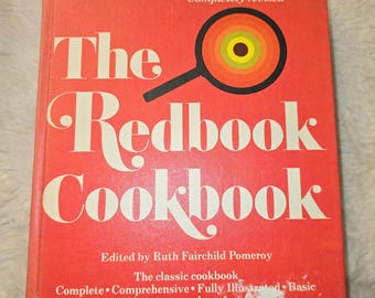May include: A hardcover book titled The Redbook Cookbook, edited by Ruth Fairchild Pomeroy. The cover is bright red with white text and a graphic of a magnifying glass with concentric circles in orange, yellow, and brown colors.