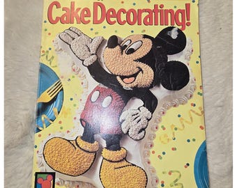 May include: The 1996 Wilton Cake Decorating! yearbook features a yellow cover with a large, textured cake shaped like Mickey Mouse. The cake is decorated with black, red, white, and yellow icing. The cover includes colorful confetti and streamers.
