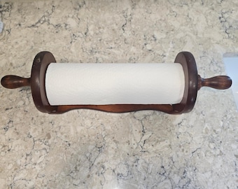 May include: A dark brown wooden paper towel holder featuring a horizontal rod with rounded handles on each end, supported by two circular side panels and a curved base, holding a roll of white paper towels in a horizontal position.