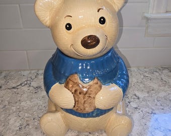May include: A ceramic cookie jar shaped like a light tan bear wearing a blue sweater. The bear has a dark brown nose, black eyes, and is holding a brown textured cookie in its paws. The surface of the jar has a glossy finish throughout.