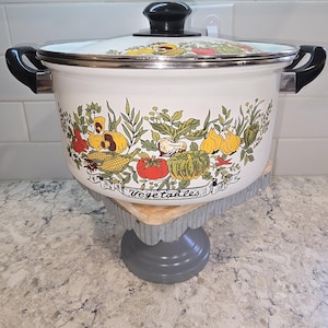 May include: A white enamel cooking pot with a black handle and lid. The pot features a colorful vegetable design around the sides, with the word "Vegetables" in a banner. The pot is sitting on a gray pedestal.
